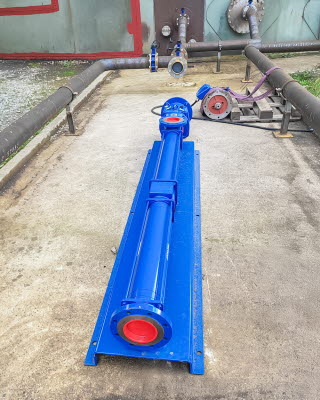 Frontansicht einer blau glänzenden Pumpe in langgestreckter Form steht auf Betonboden, dahinter Rohrleitungen und ein Stück Wiese