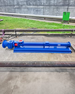 Seitenansicht einer blau glänzenden Pumpe in langgestreckter Form steht auf Betonboden, dahinter Rohrleitungen und ein Stück Wiese